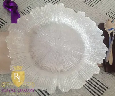 Pearl white textured glass charger with ruffled rim, set on a striped table beside a folded napkin and spoon