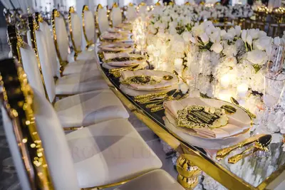Close-up of a place setting on a curved gold table with mirror top, white roses and gold cutlery, white chairs alongside