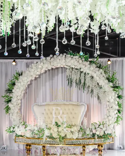 Gold and crystal glass sweetheart table styled at a wedding, beneath a white floral ring, a white loveseat and hanging