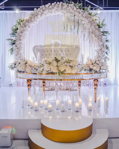Curved gold table with crystal edge on a white stage, white flowers above, pillar candles on the floor, floral ring behind