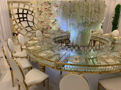 Large round table assembled from gold-framed glass sections, set with plates and glassware beside white hanging wisteria