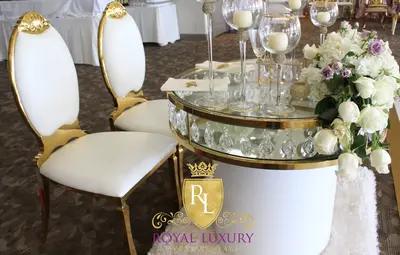 Gold-edged glass and crystal table with white base beside white gold-framed chairs, topped with candle holders and white