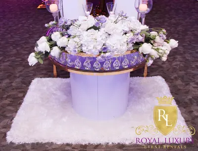 Round table with white base on a white shaggy rug, its glass top covered in white and lilac roses