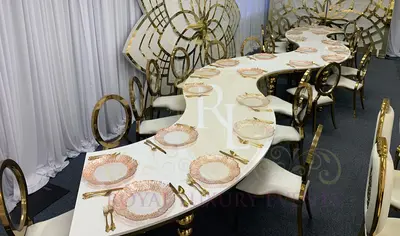 Serpentine table with white top and gold frame, set with pink-toned plates, gold cutlery and gold ring-back chairs
