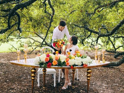 Couple at a curved white table with gold bubble legs, orange and pink roses and gold candleholders under an oak tree