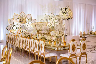 Long gold and crystal dining table beside white and gold chairs, styled before a gold floral backdrop at a wedding