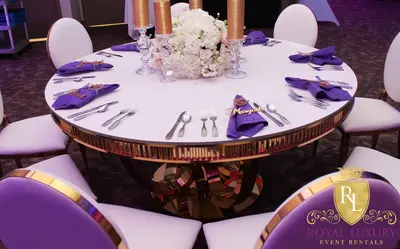 Round white-topped gold table set for dining with purple napkins, white and purple chairs and a white floral centrepiece
