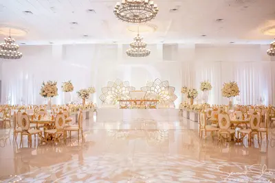 White draped ballroom wall behind a gold lotus-shaped backdrop, with tall white floral arrangements and gold chairs