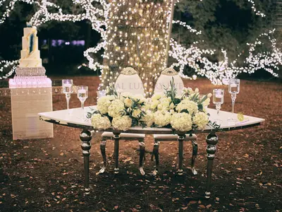 Curved white-topped sweetheart table with silver bubble legs, white hydrangea centrepiece and two chairs, set beneath a lit