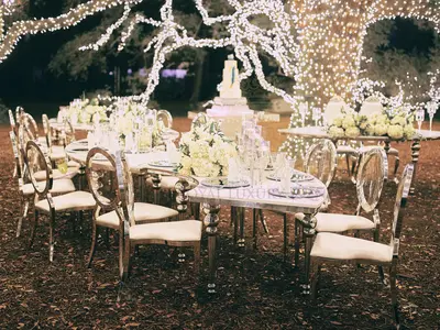 Serpentine white-topped tables with silver bubble legs set for dinner among oval-back chairs, white florals and a lit tree