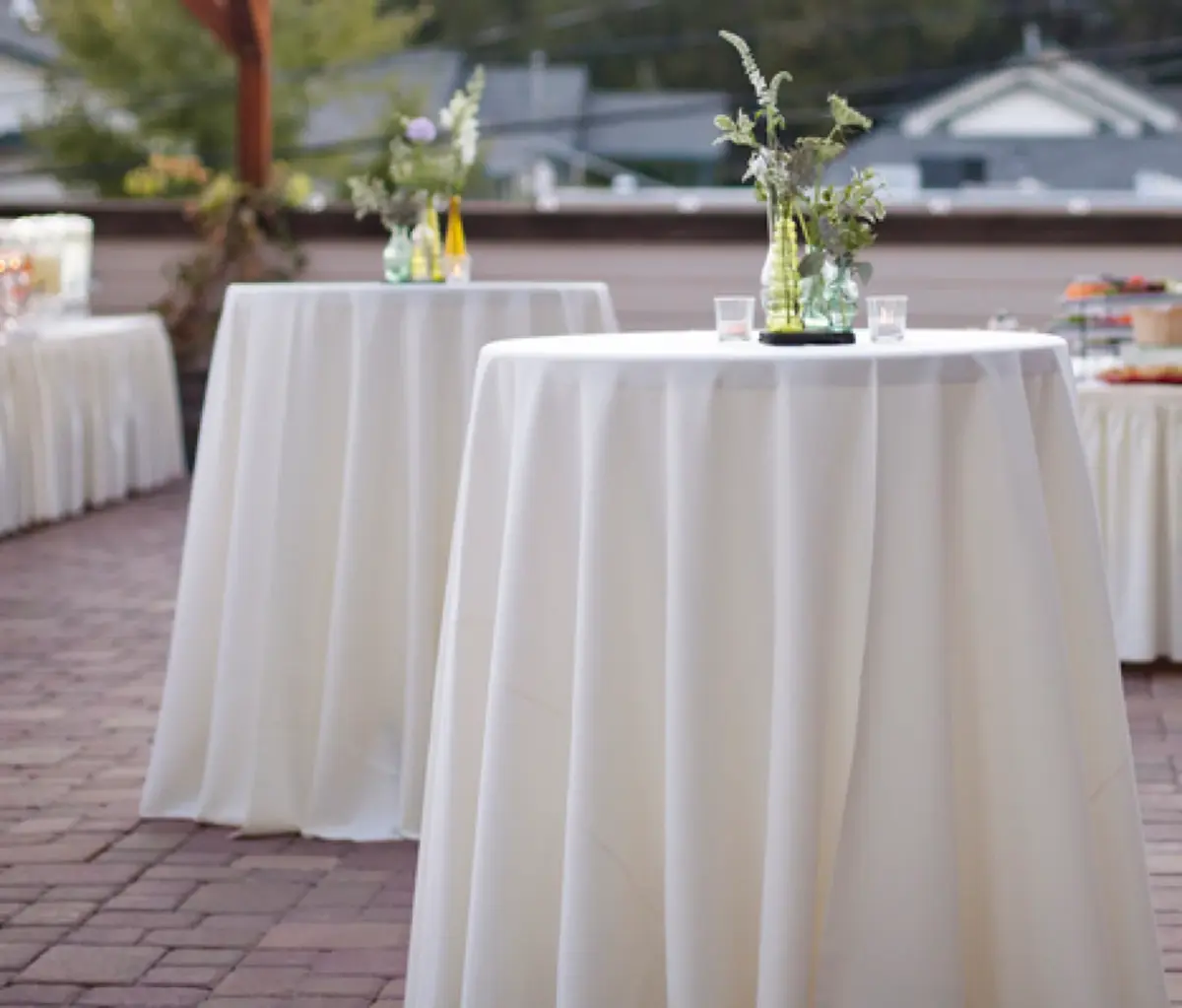Round white cocktail tablecloths with soft drape on bistro tables outdoors, with small vases of greenery on a brick patio