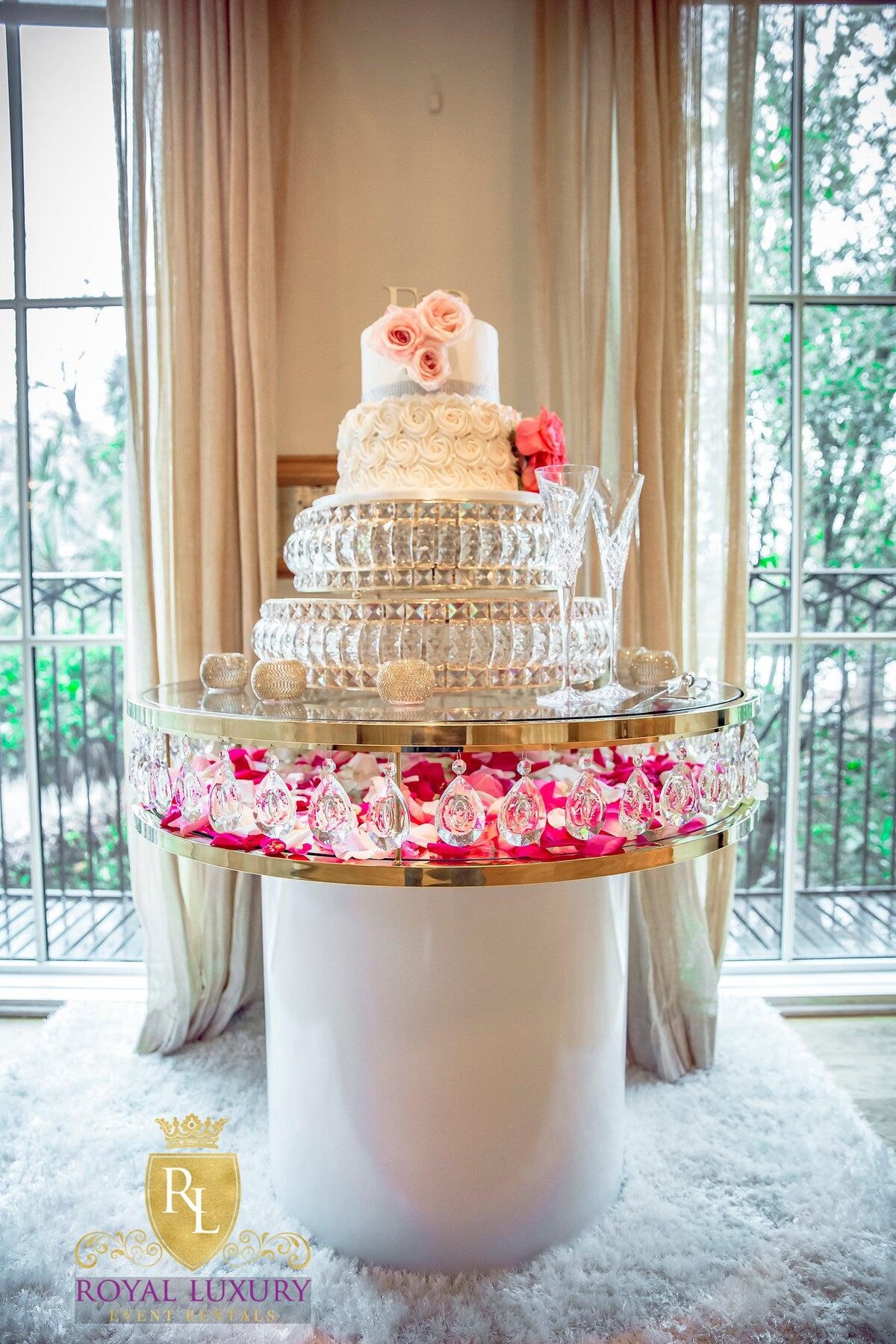 Round gold-rimmed glass cake table with white base, crystal drops and pink petals, holding a tiered rose cake