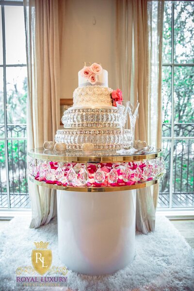 Round gold-rimmed glass cake table with white base, crystal drops and pink petals, holding a tiered rose cake