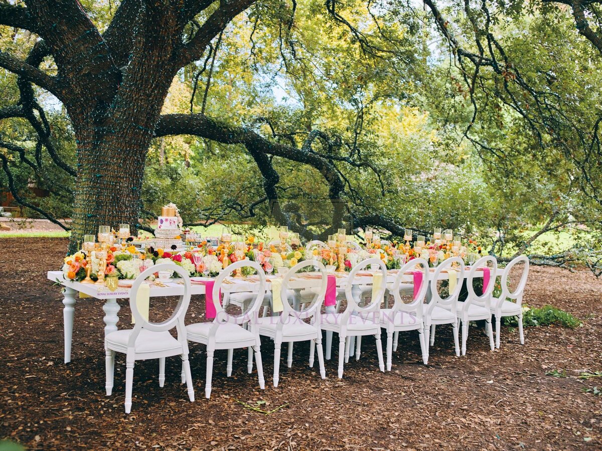 White rectangular dining table with turned legs under an oak tree, set with colourful flowers and white oval-back chairs