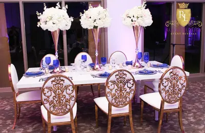 White high-gloss table styled indoors with tall white floral centrepieces, blue glassware and gold damask-back chairs