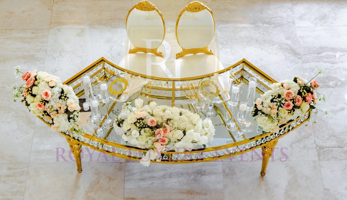 Curved gold sweetheart table seen from above, glass top with crystal edge, set with pink and white floral arrangements