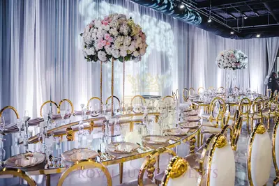 Gold-framed mirror-top table with tall pink and white floral arrangements, styled at a reception against white draping