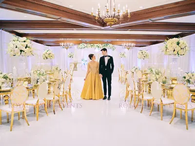 Couple standing in a ballroom between rows of gold-framed tables and ivory chairs with white floral centrepieces