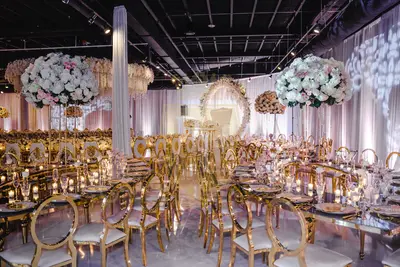 Mirror-topped tables and gold-framed chairs at a wedding reception, with tall white hydrangea centerpieces and a floral arch