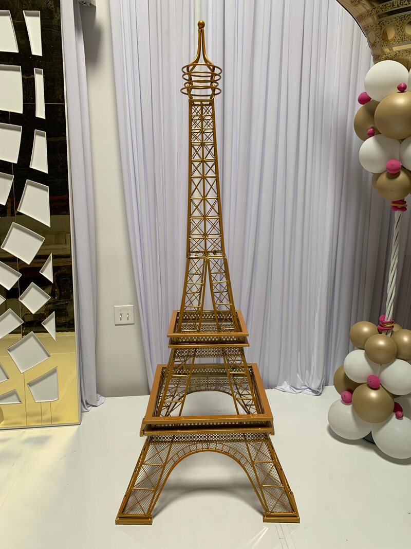 Gold lattice Eiffel Tower replica standing on a white floor before white draping, beside gold and white balloons