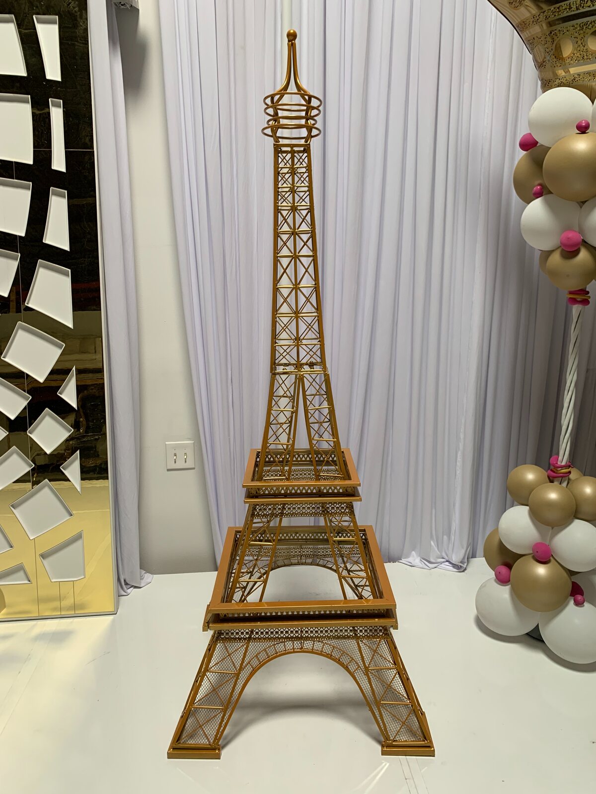 Gold lattice Eiffel Tower replica standing on a white floor before white draping, beside gold and white balloons