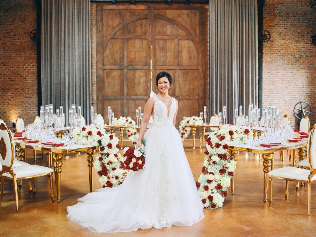 luxurious indoor wedding reception decor design, with red, and white floral designs, gold and white curved tables, gold chairs, and a stunning bride and bridal bouquet.