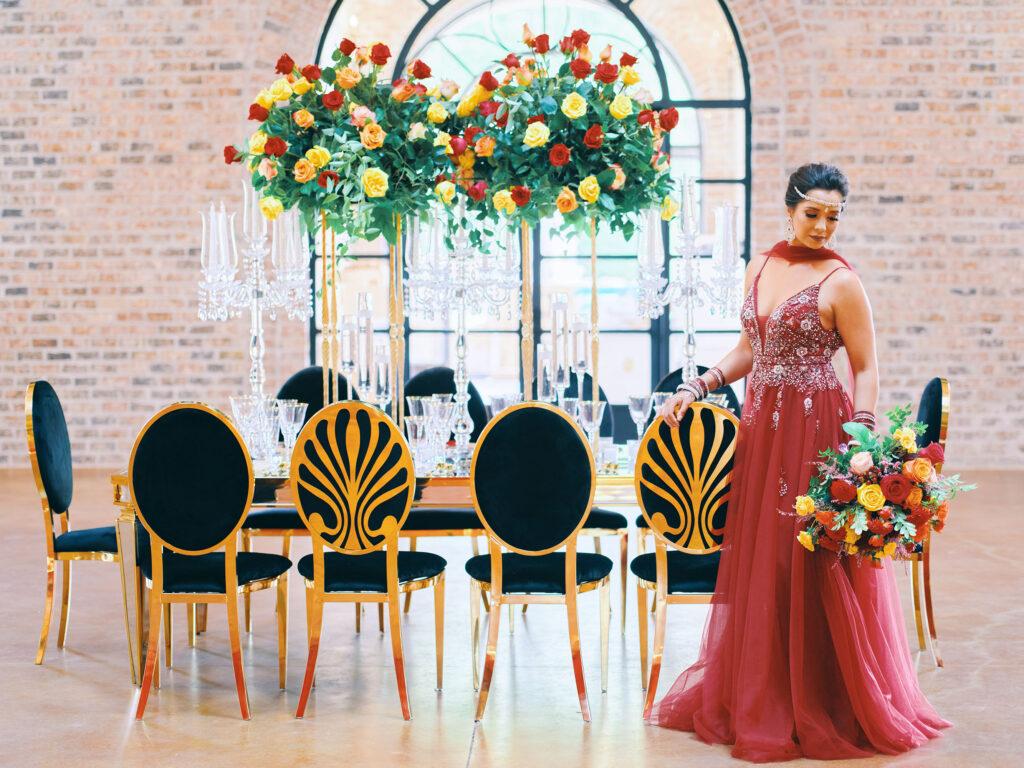 luxurious indoor wedding reception decor design, with red, yellow and orange floral designs, gold and black chairs, rectangular table, and a stunning bride and bridal bouquet.