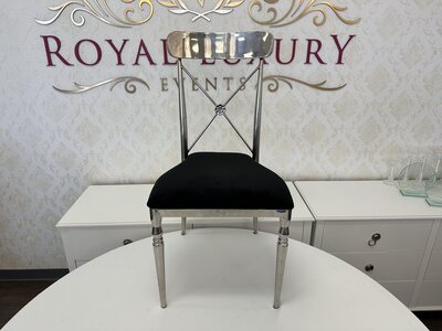Silver cross-back dining chair with black velvet seat, shown front-on in a showroom with a damask wall behind