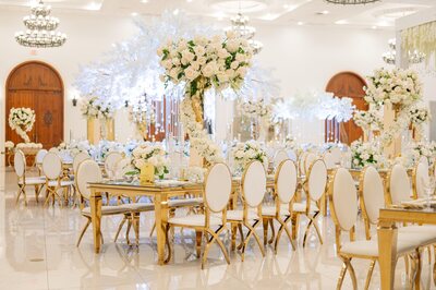 Gold mirror-top dining tables with white and gold oval-back chairs, styled at a reception with white floral centerpieces
