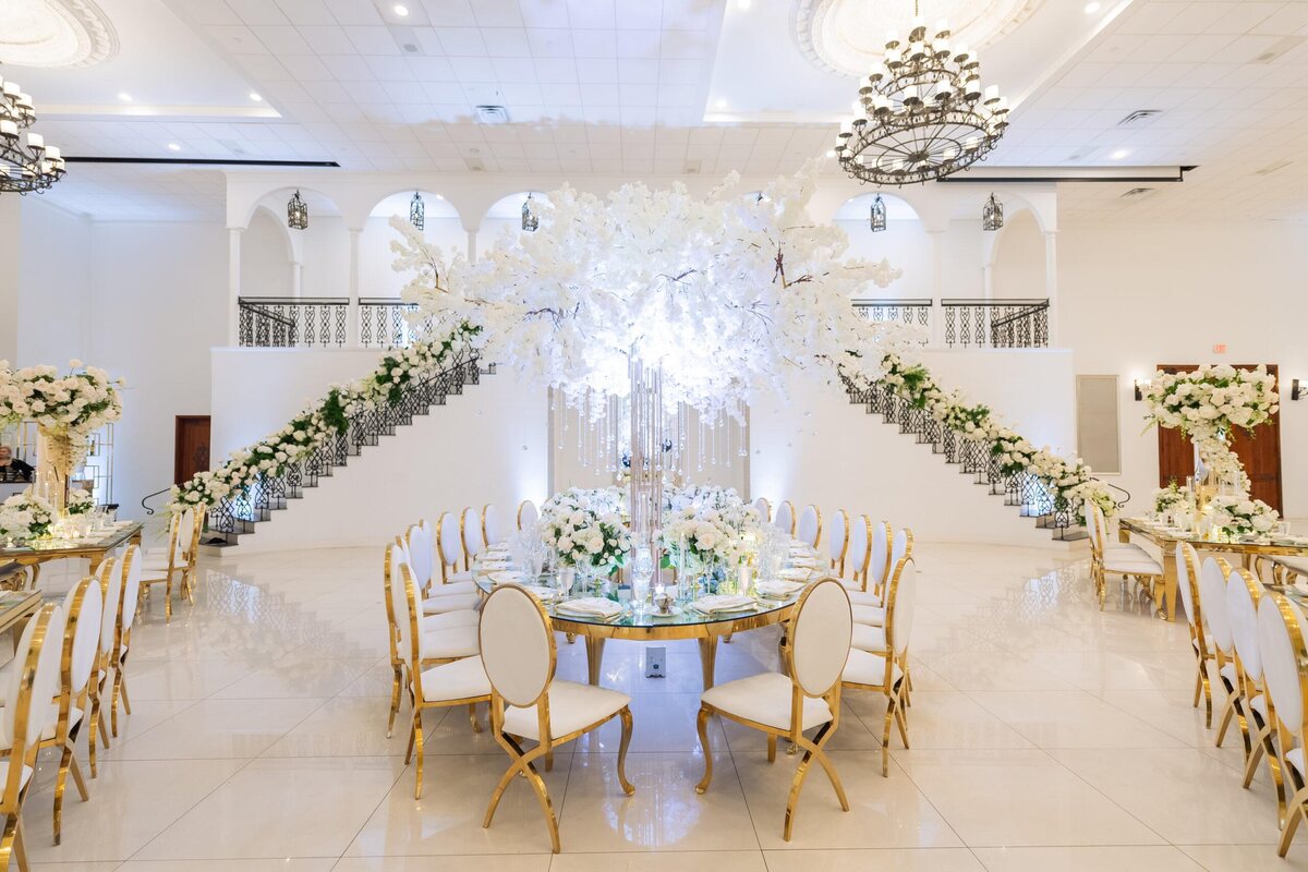Large gold oval mirror-top dining table styled at a wedding reception, with a white blossom centerpiece and gold chairs