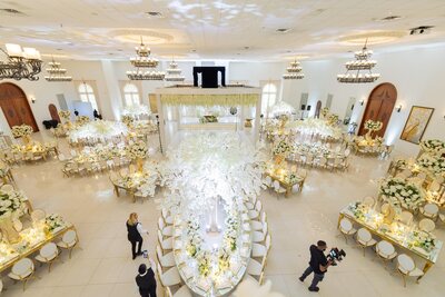 Overhead view of a ballroom wedding with a long gold oval table under a white blossom tree, other tables beyond