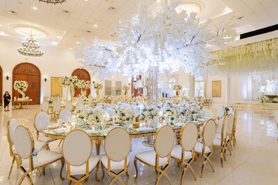Gold oval table with mirror top set with white florals and glassware, surrounded by white and gold oval-back chairs