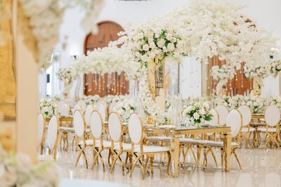 Gold dining table with mirror top and white floral arrangements, seen from a low angle among white-cushioned gold chairs