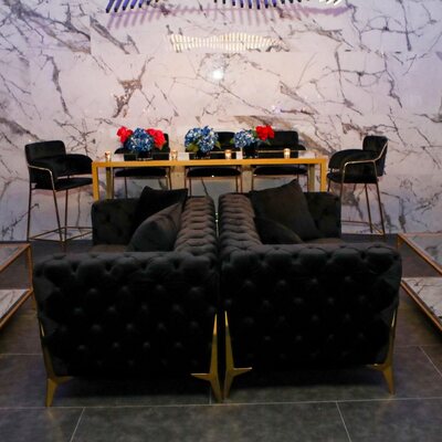 Two black tufted velvet sofas with gold legs seen from behind, set before a gold bar table at a marble-walled event