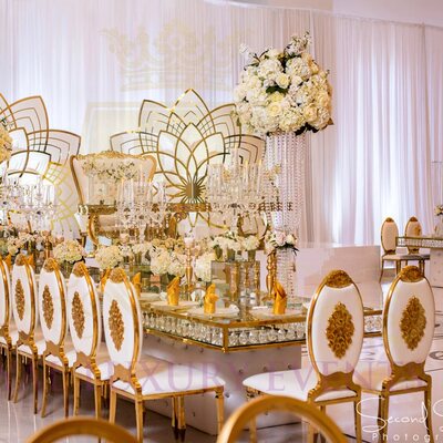 Long head table lined with gold chairs bearing white cushions and gold medallions, beneath white and gold lotus-shaped