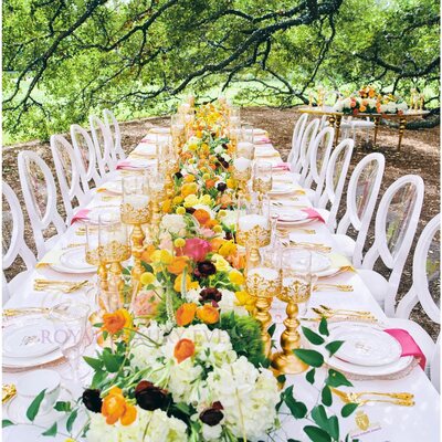 Rows of white clear-back Louis chairs along a long white banquet table with gold candle stands and orange floral runner