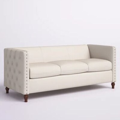 Beige three-seat lounge sofa with low back, tufted side panel, nailhead trim and dark turned wooden legs, seen at an angle