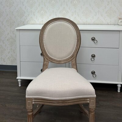 Front view of a cream-upholstered Louis chair with oval back, set before a white dresser in a showroom