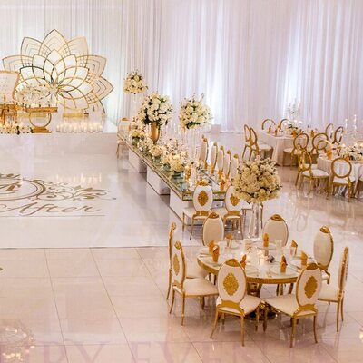 Ballroom wedding reception with gold and white chairs at round tables, white draping and a gold floral backdrop behind the