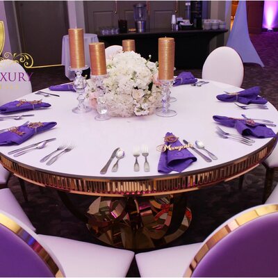 Round table with white oval-back gold chairs, purple napkins, gold candles and white floral centerpiece at a reception