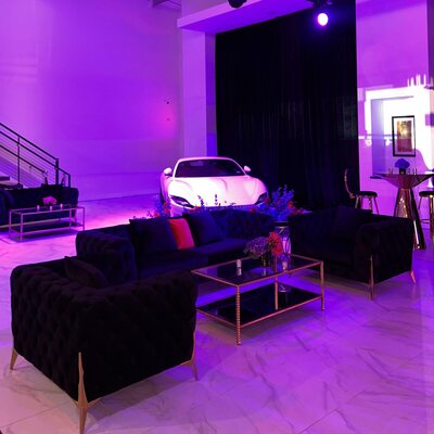 Black tufted velvet sofa and armchairs around a gold-framed glass coffee table, styled near a white sports car at an event