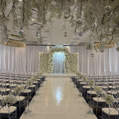 Clear lattice-back chairs with white cushions in rows beside a white aisle, styled at a ceremony with flowers and crystal