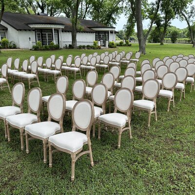 Rows of wooden Louis chairs with cream cushioned seats set on a lawn for an outdoor ceremony beside a white house