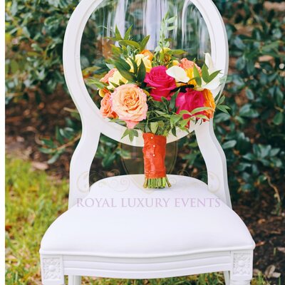 White Louis chair with clear oval back outdoors on grass, holding a coral and pink rose bouquet on the seat