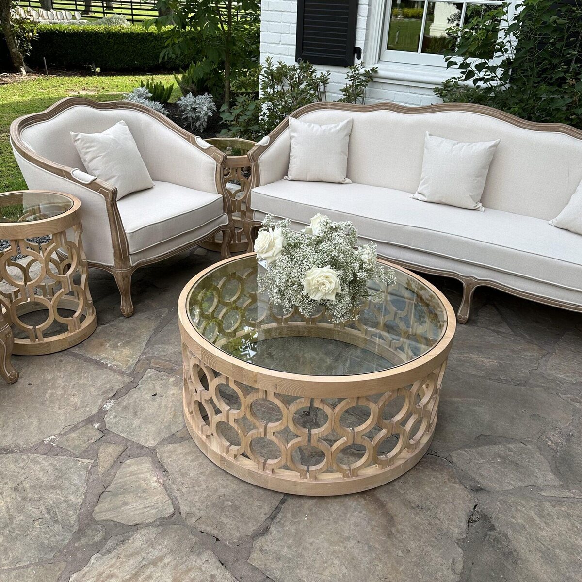 Off-white lounge chair with natural wood frame beside a matching sofa and round coffee table on a stone patio